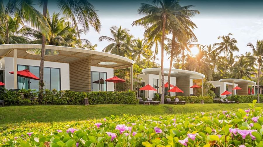Row of modern white villas with large glass windows, red umbrellas, and sun loungers surrounded by palm trees and flowering greenery at Ramada Resort by Wyndham Khao Lak.