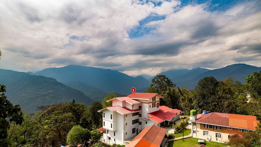 Aerial view of a hillside resort with white and red-roofed buildings surrounded by lush greenery, set against a backdrop of mountains and a dramatic cloudy sky - Yashshree Pelling Chekhim Retreat.