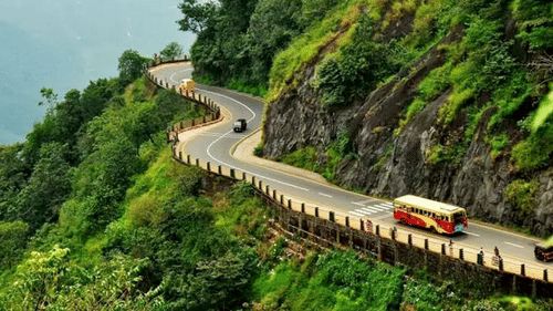 Vehicles drive through a winding road with a hill face on one side and a drop on the other, as seen from Lakkidi View Point in Wayanad.