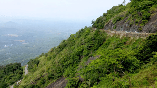A winding mountain road carved into a steep, lush green cliff side overlooking a vast, misty valley.