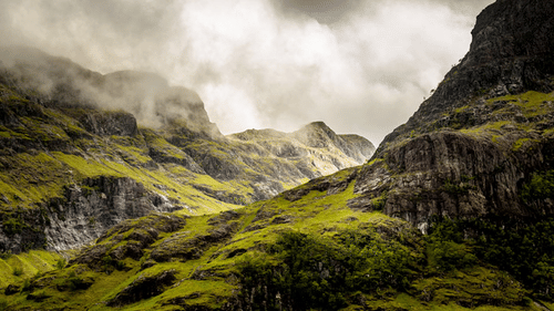 A landscape of steep, rocky mountains covered in green grass, with low-hanging clouds obscuring the peaks.