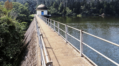 A dam with a walkway along the side, surrounded by dense forested hills and water below.