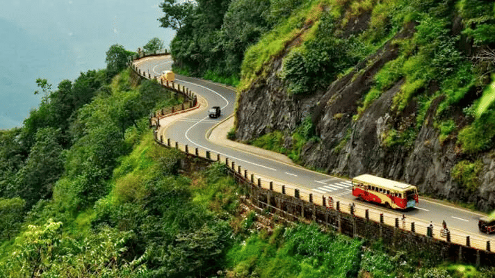 Vehicles drive through a winding road with a hill face on one side and a drop on the other, as seen from Lakkidi View Point in Wayanad.