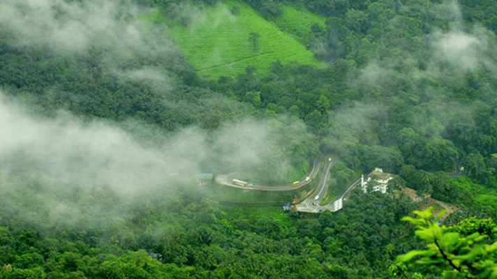 Aerial view of a winding mountain road with vehicles driving through them, surrounded by greenery on either side and mist rising above it.