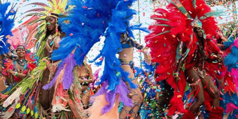 A lively group of revelers in large, brightly feathered carnival costumes dance with confetti falling around them.