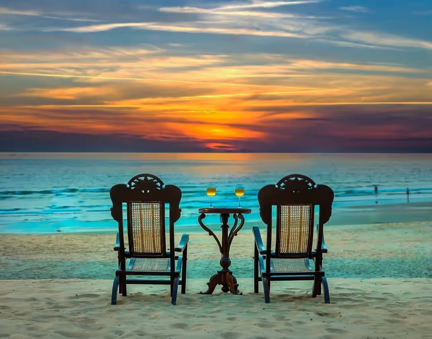 two chairs and a table with two glasses kept on it in front of a sun set - Caravela Beach Resort Goa
