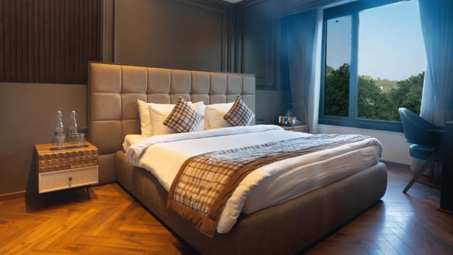 A bedroom featuring a bed with white linens, patterned runner, wooden flooring, and a large window overlooking green trees | Lawrence Solitaire Resort, Nainital