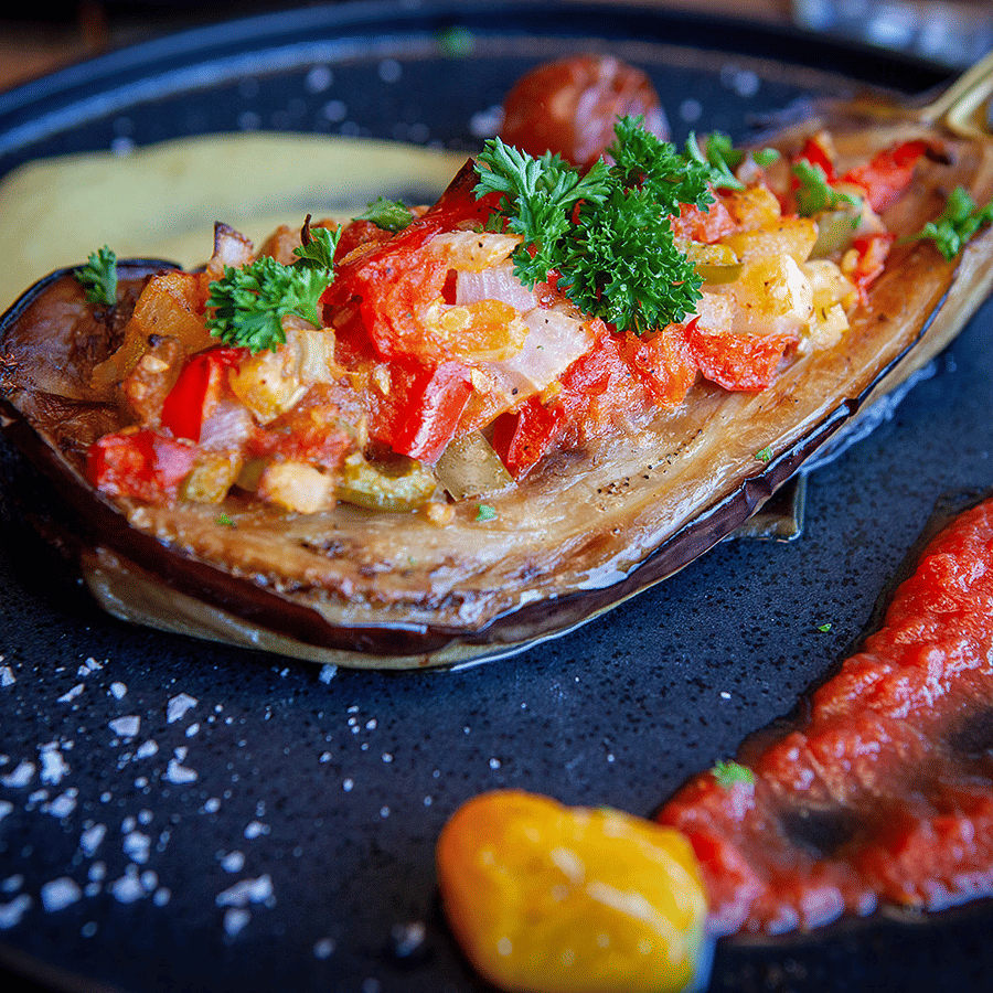 A roasted eggplant topped with diced tomatoes, peppers and fresh parsley, served with sauces on a dark plate at YO1 Longevity & Health Resorts