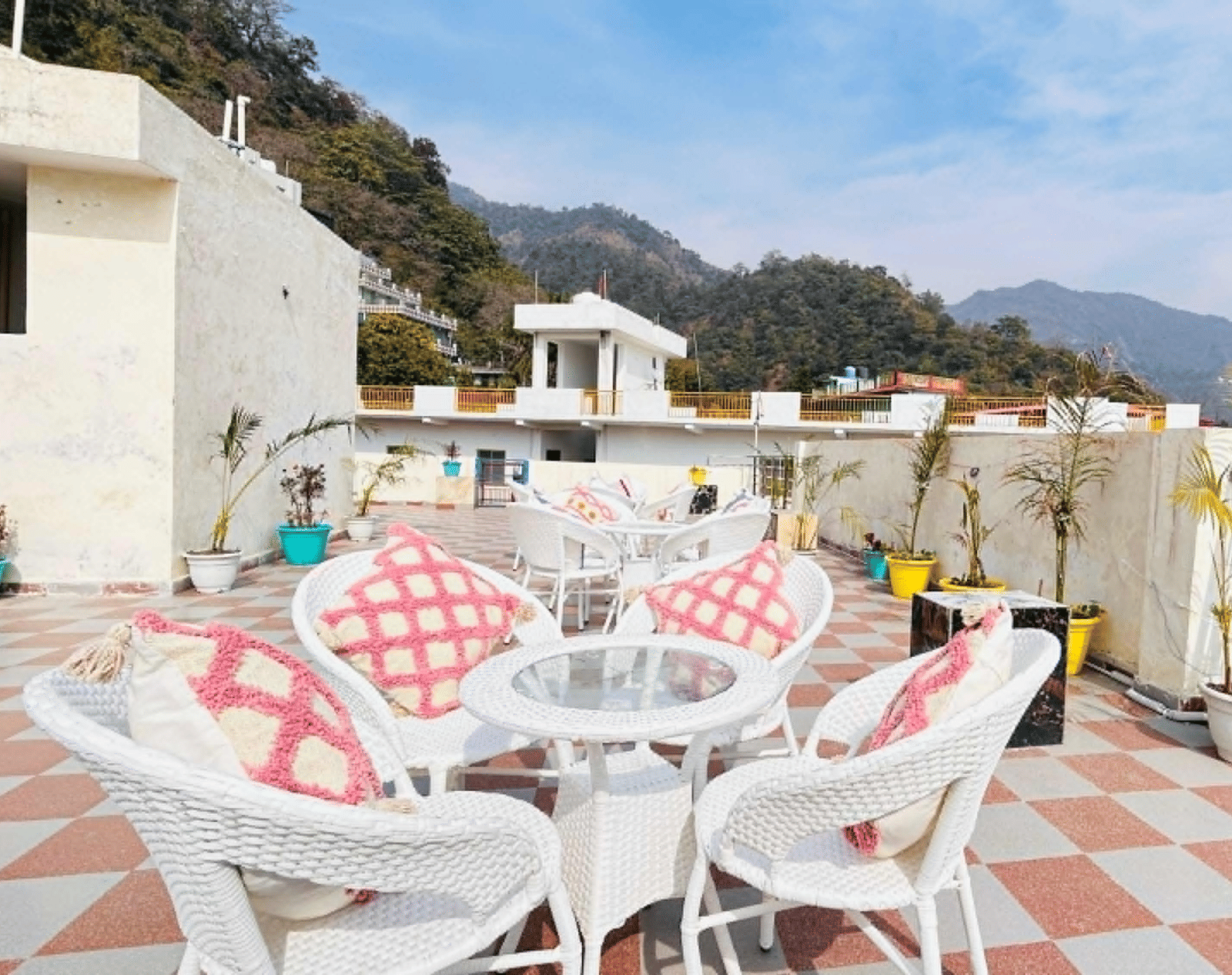 An outdoor rooftop terrace with white wicker tables and chairs featuring a red and white checkered pattern, set against a background of green hills | Perfectstayz Value Hills