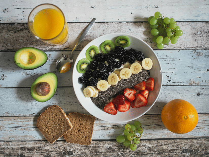 A bowl of yogurt topped with kiwi, blackberries, banana, strawberries and chia seeds beside toast, avocado, grapes, an orange and a glass of juice