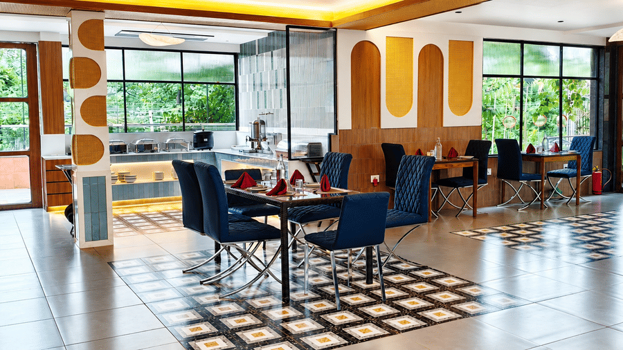 Spacious dining hall with large glass windows, upholstered dining chairs, decorative wall panels, and ample natural light at Kali Mirch Restaurant, Lords Riverside Resort, Muduba.