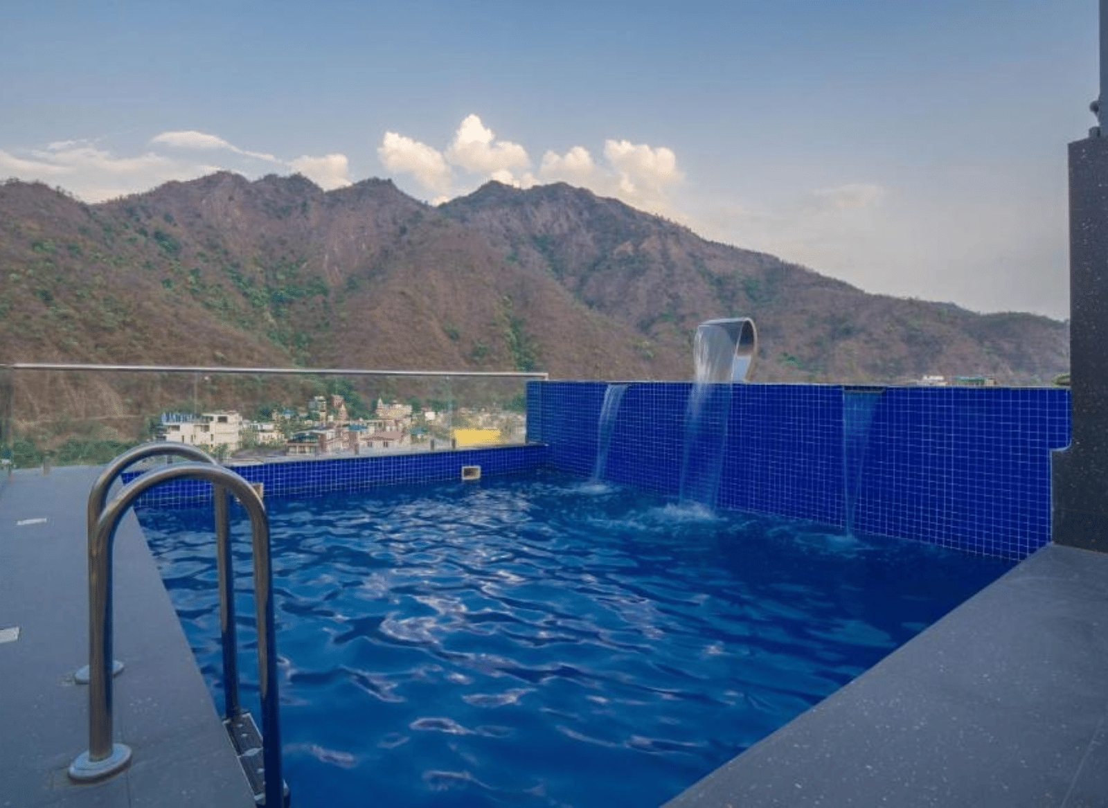 An outdoor swimming pool with metal handrails, water flowing from a spout, and a mountain range visible in the background under a clear sky at Perfectstayz Premium Laxmi Heritage.