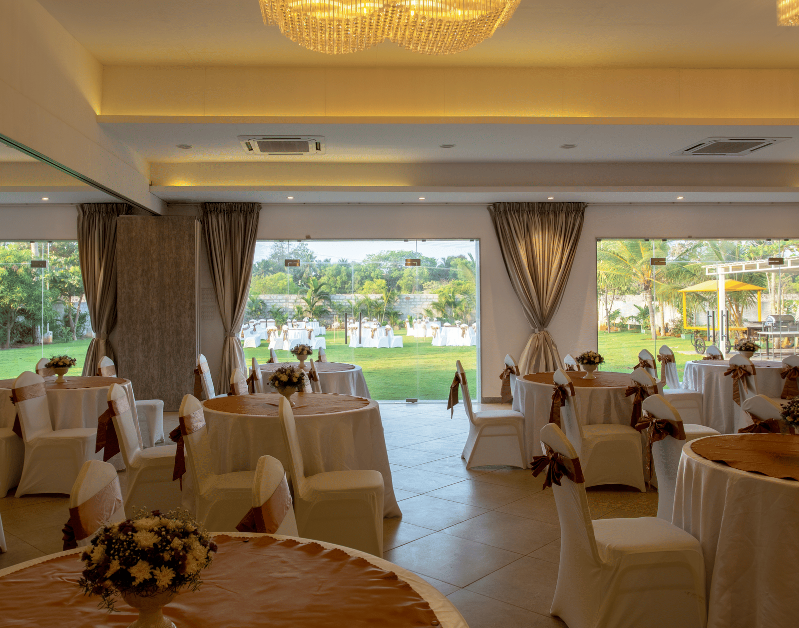 An indoor banquet hall with round tables and chairs draped in white with brown sashes, set for an event - Grande Bay Resort & Spa, Mamallapuram
