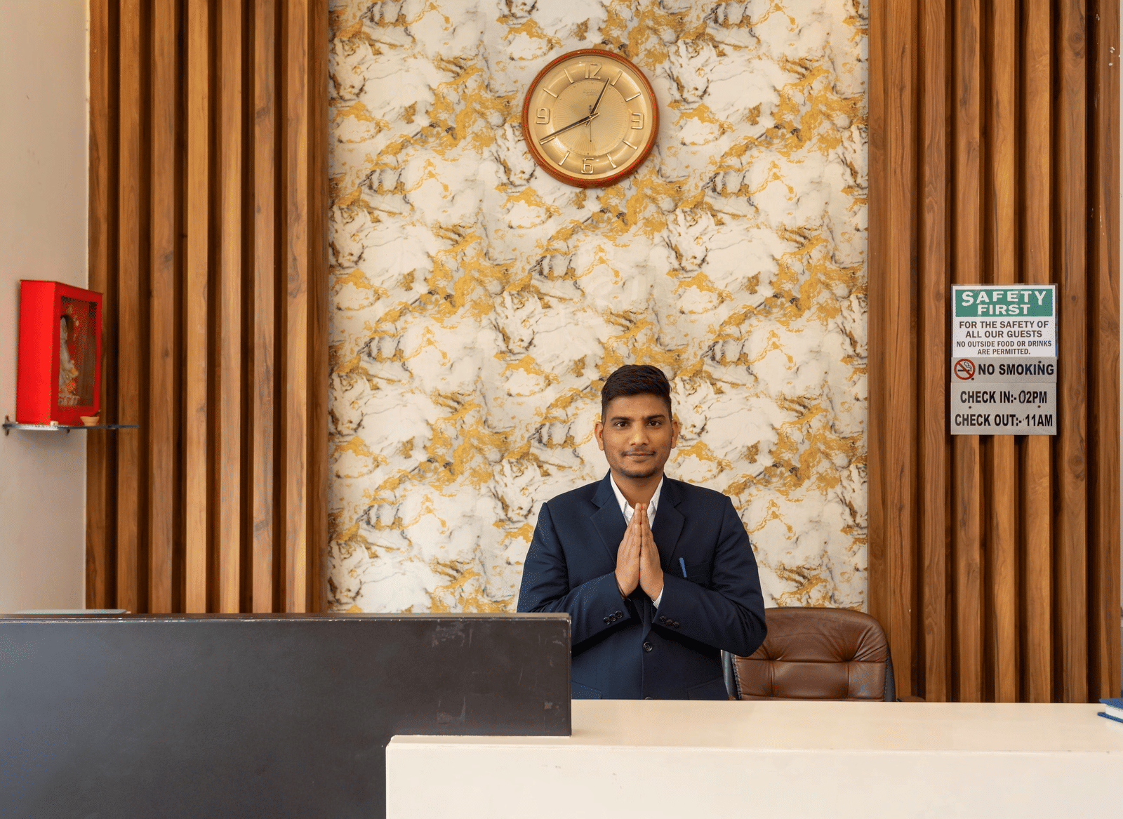 A man standing behind a reception desk with a wall clock and textured wallpaper at Perfectstayz Premium Shanti Heritage, Haridwar