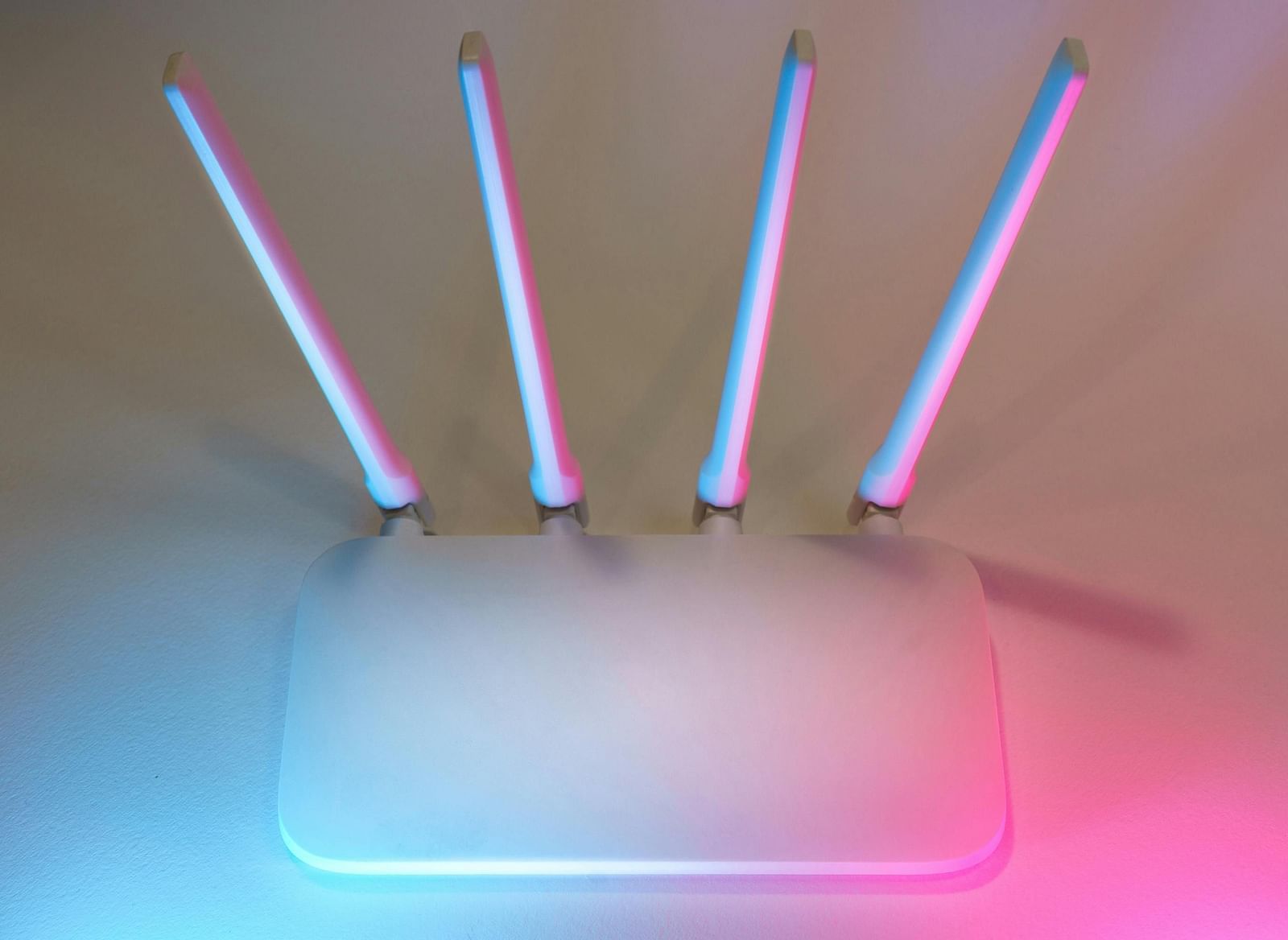 A white wireless router with four antennae illuminated in blue and pink hues against a neutral wall.