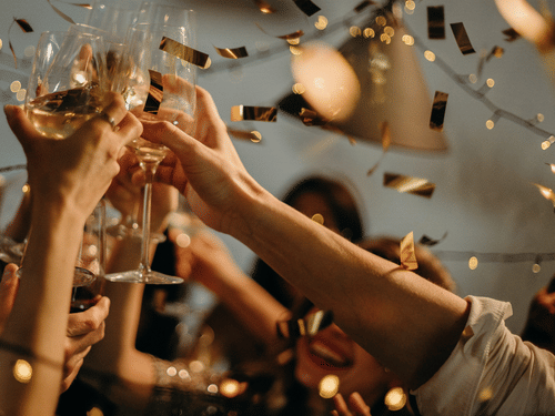 People raising champagne glasses in a toast during a celebration with falling gold confetti and fairy lights.