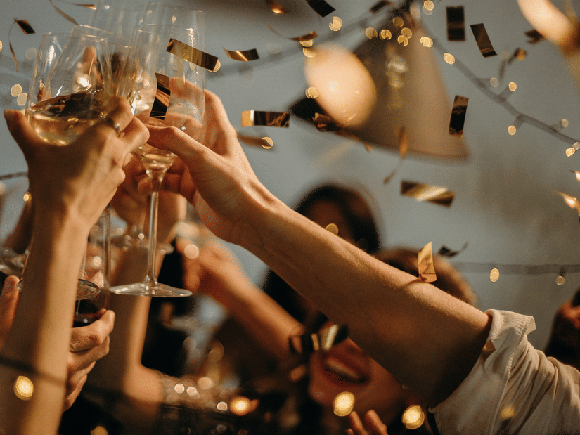 People raising champagne glasses in a toast during a celebration with falling gold confetti and fairy lights.