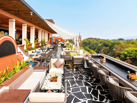 Rows of dining tables, chairs, and outdoor lounge seating line a spacious open terrace at The Sierra - By The Lake, next to a lush green forest view.