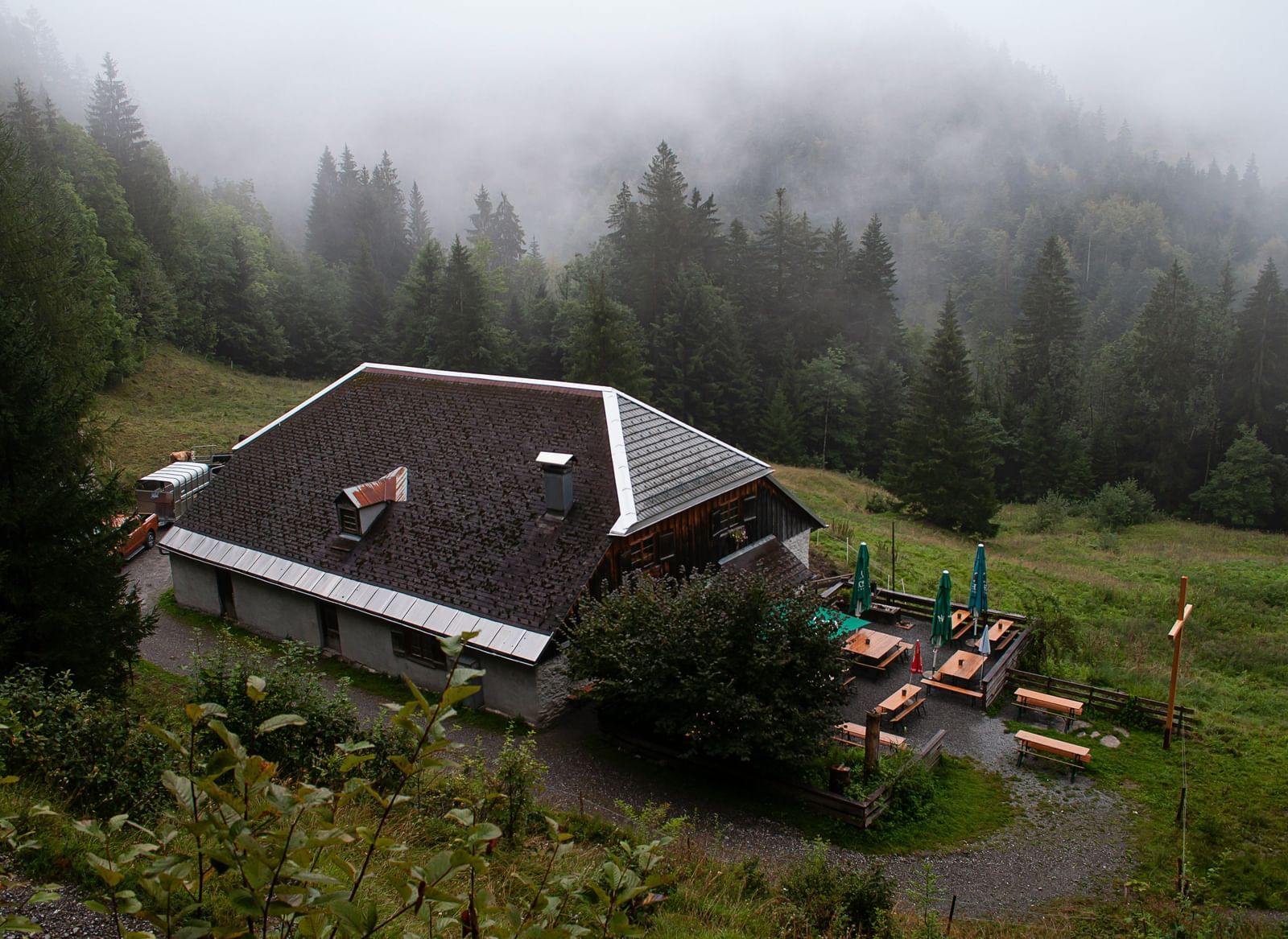 An isolated wooden house with a dark roof nestled on a forested hillside, surrounded by mist or fog.
