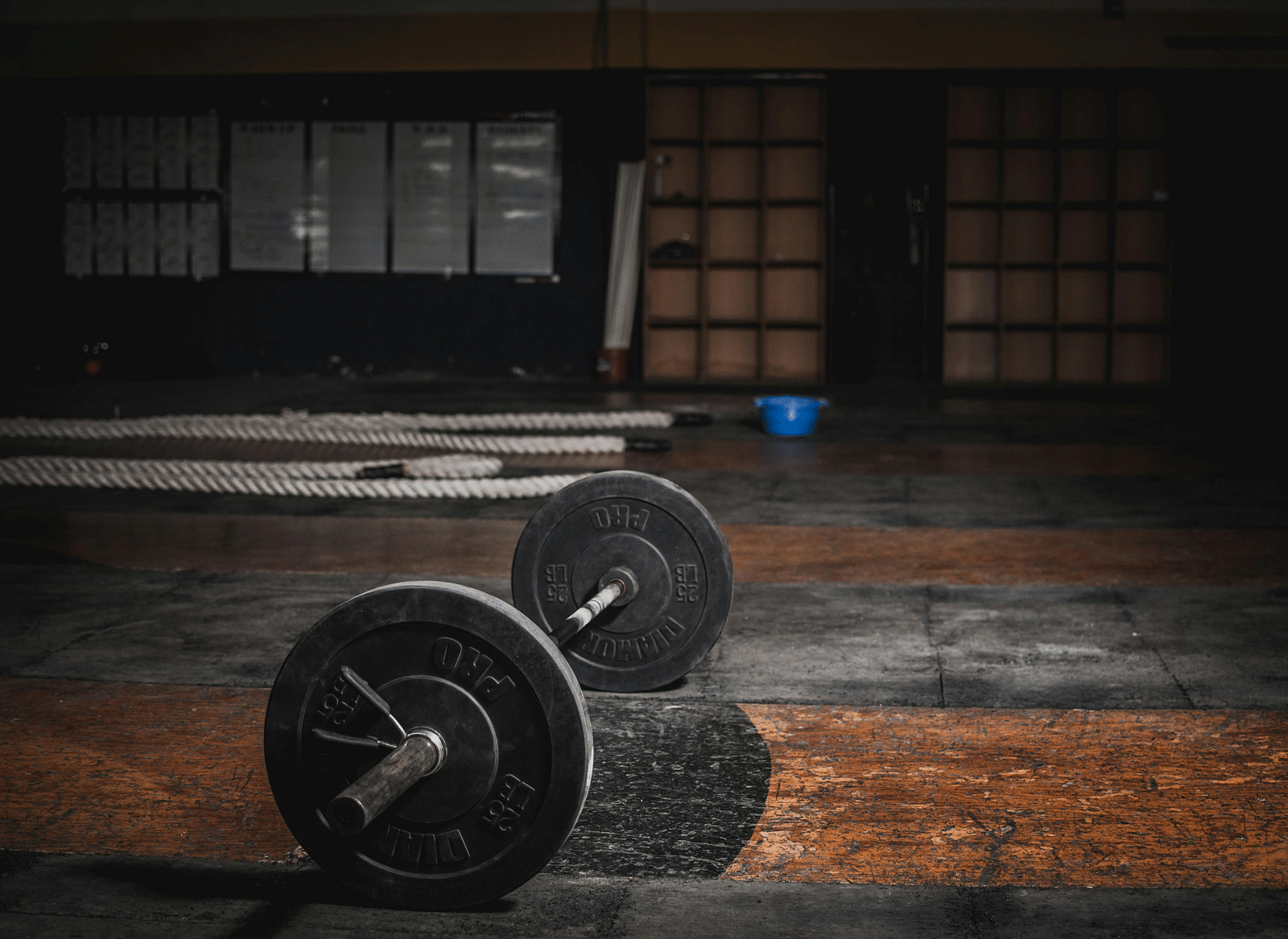 A barbell with weights lies on a worn gym floor, with long metal bars in the background.