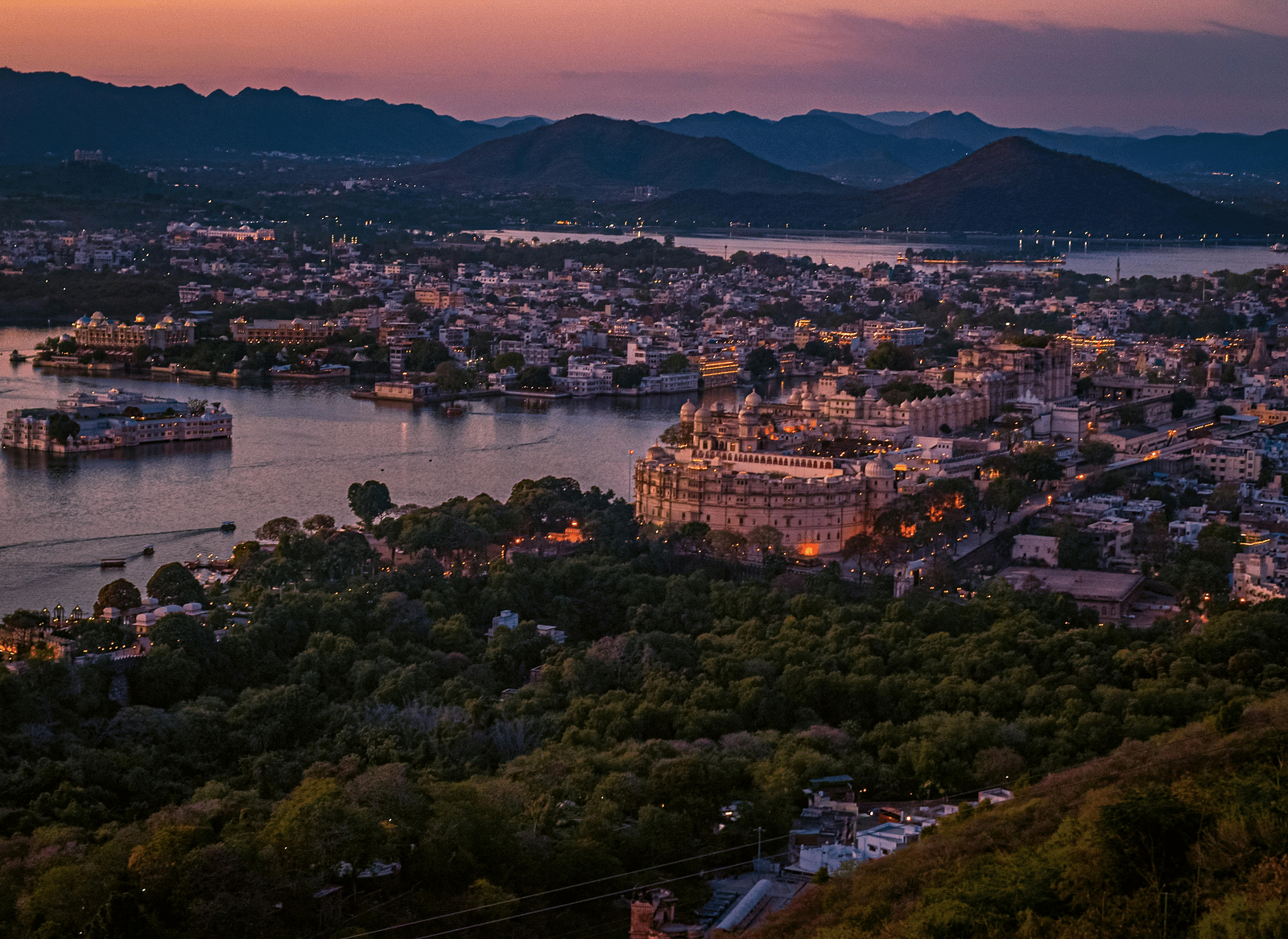 A twilight aerial view captures Udaipur, India, showcasing the City Palace lit with warm lights, nestled beside Lake Pichola, surrounded by a densely populated city and distant hills.