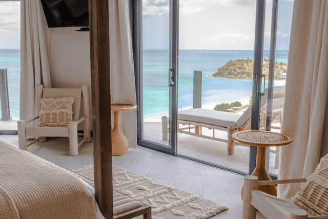 A bright, airy bedroom with a large bed and a view of the ocean through an open doorway, suggesting a beachfront resort.