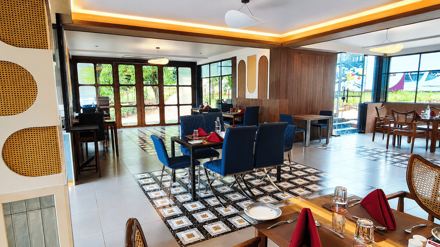 Sunlit dining area with upholstered chairs, wooden tables, large glass windows, and patterned flooring at Kali Mirch Restaurant, Lords Riverside Resort, Muduba.