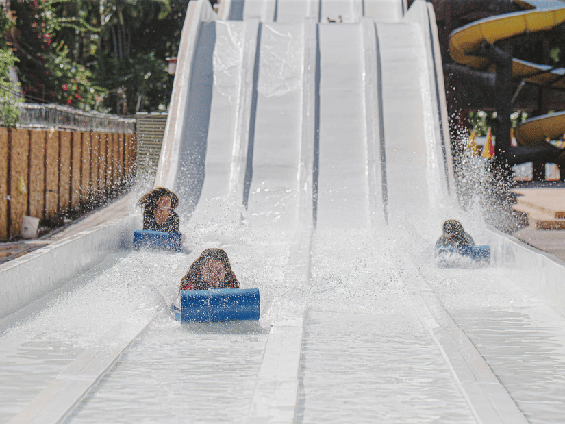 A view of three children riding a mat racer slide at a water park with trees on the left hand side and a swirling slide on the right