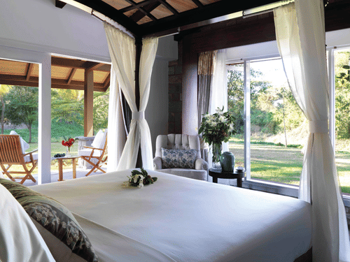 A bright, airy bedroom at The Residence, The Serai Kabini, with a canopy bed and sheer curtains, large windows opening to lush greenery, an armchair, and a covered patio with wooden chairs overlooking the garden for a relaxing stay in Kabini.