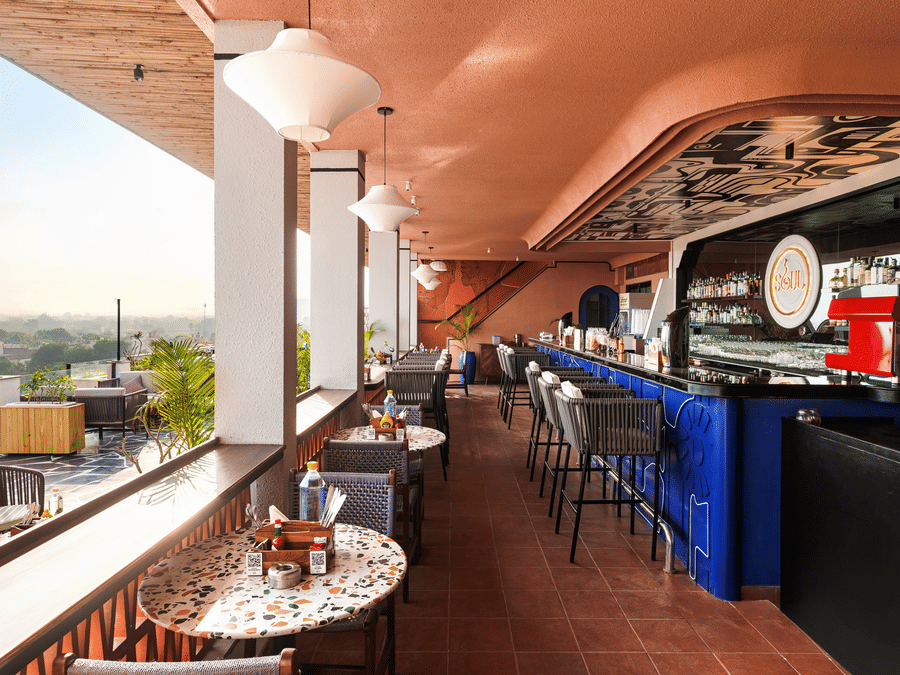 Stools line a vibrant bar counter on a rooftop patio at The Sierra - By The Lake, showing a view of the surrounding landscape past the open railing.