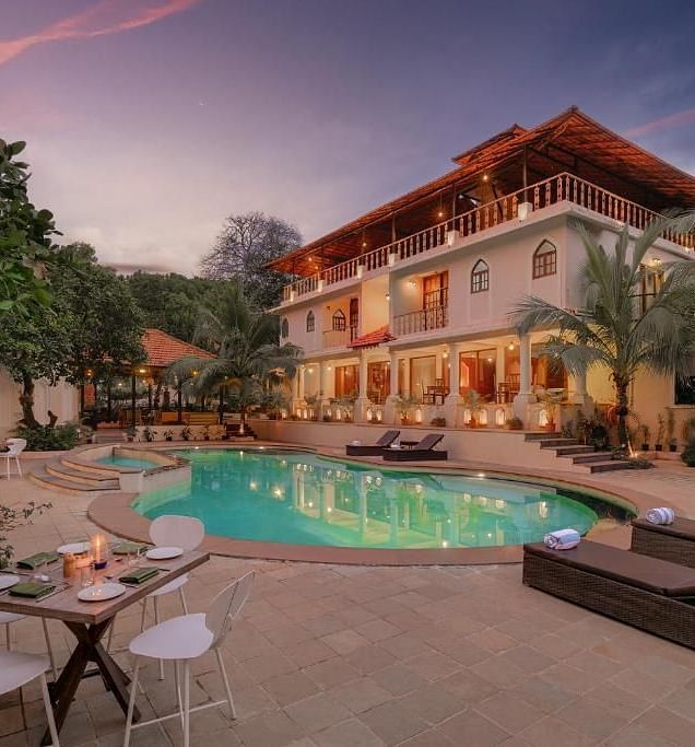 An image of Amritara Aura featuring a well lit facade, a swimming pool in front of it as well as outdoor seating area