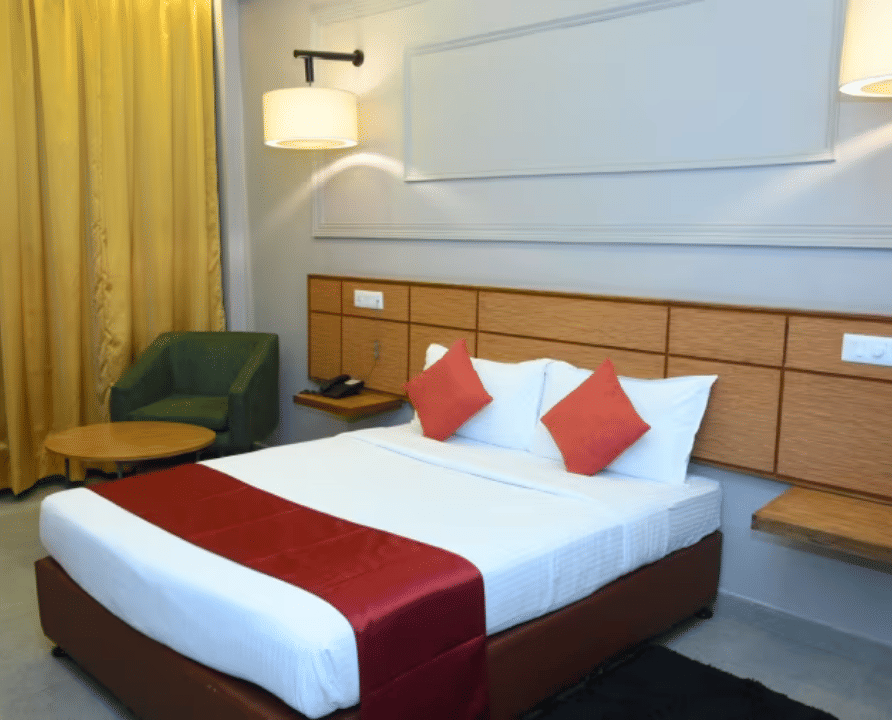 Bedroom with double bed, red runner, pillows, bedside lamps and yellow curtains at Max Hotel Prayagraj.