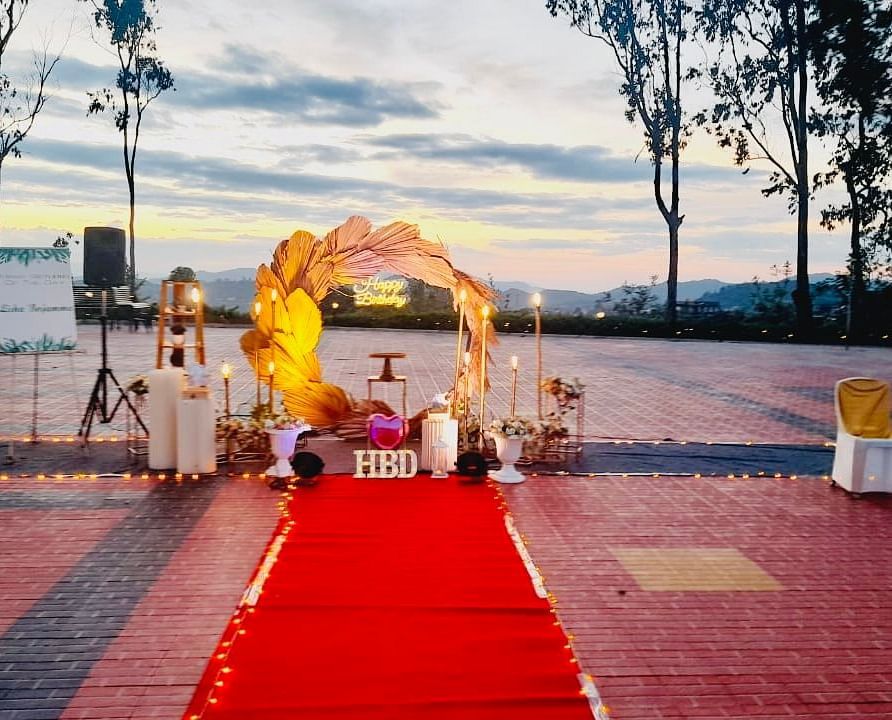 An outdoor wedding setup features a red carpet leading to a floral archway, with chairs arranged on either side and greenery in the background | Open Deck | Woodstock Resorts, Coorg