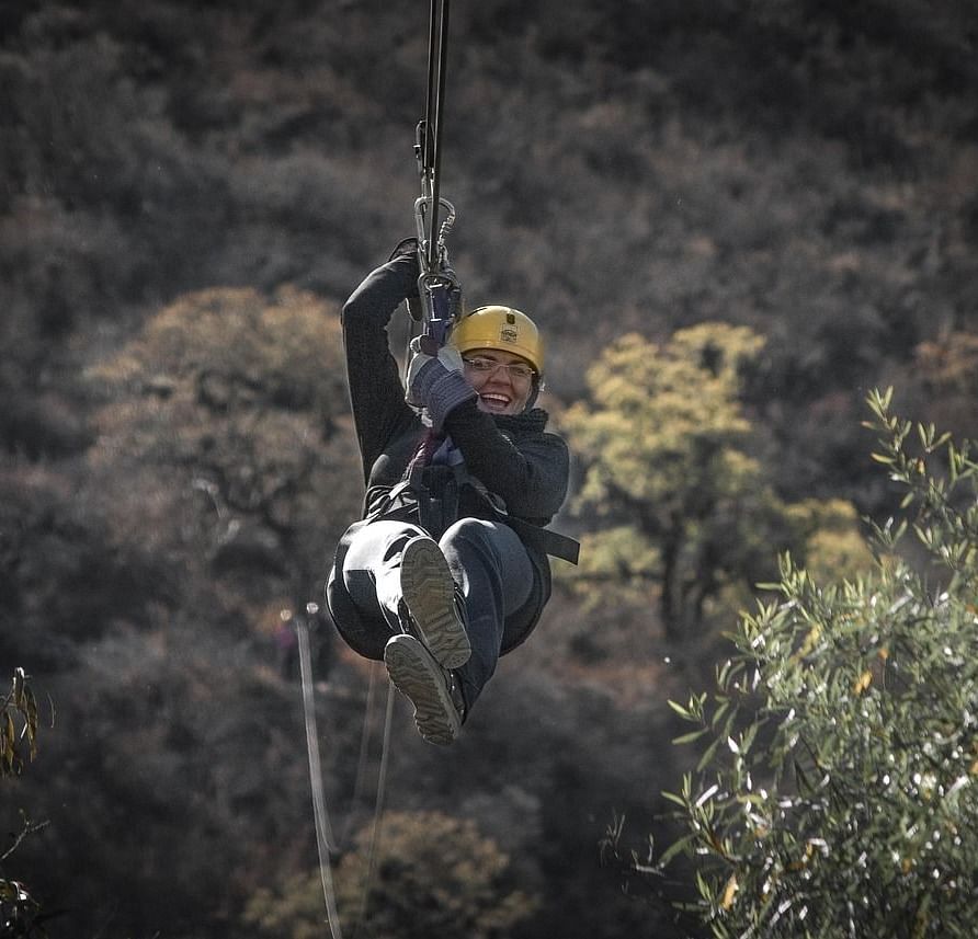 A person holding on to the zip line wearing safety gear with a mountain terrain in the background.