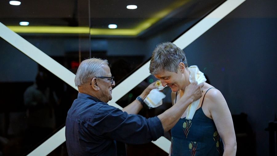 A man with glasses adjusts a white garment around a woman's neck in a salon or similar setting.