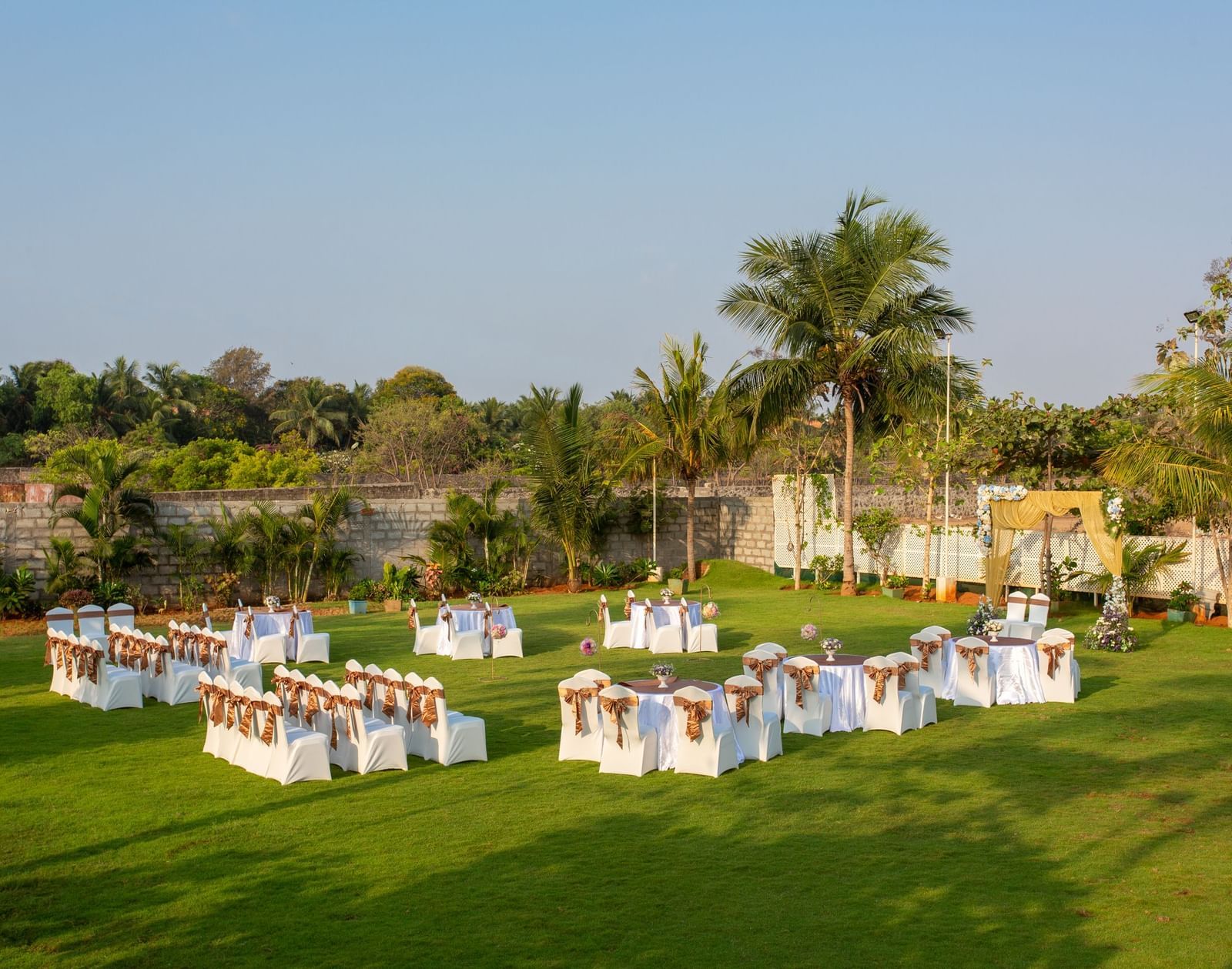 An outdoor event setup on a green lawn with tables and chairs draped in white and yellow, with palm trees under a bright sky | Grande Pavillion | Grand Bay Resort and Spa