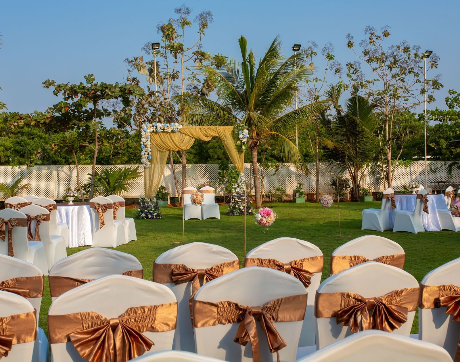 An outdoor event space with chairs covered in gold sashes, arranged on a green lawn, with a decorative archway in the distance | Grande Pavillion | Grand Bay Resort and Spa