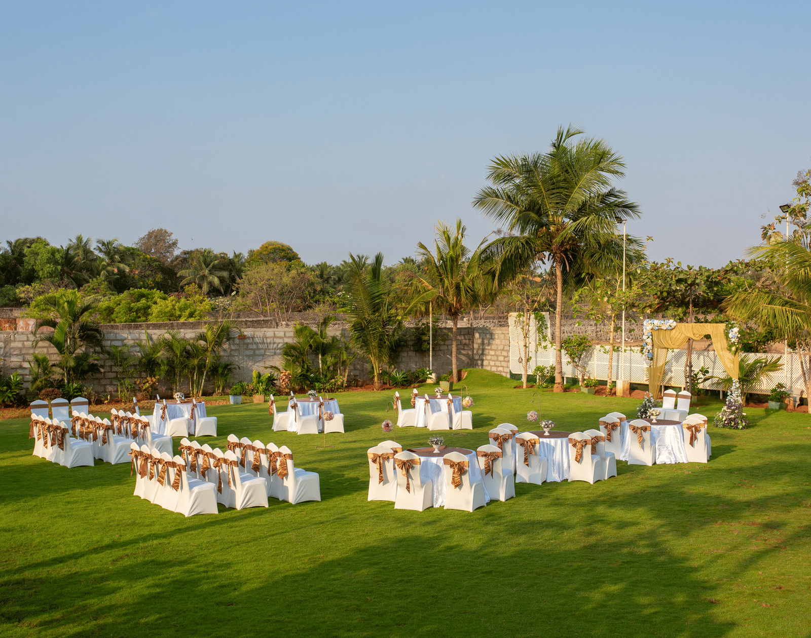 An outdoor event setup on a green lawn with tables and chairs draped in white and yellow, surrounded by coconut trees and under a bright sky - Grande Bay Resort & Spa, Mamallapuram