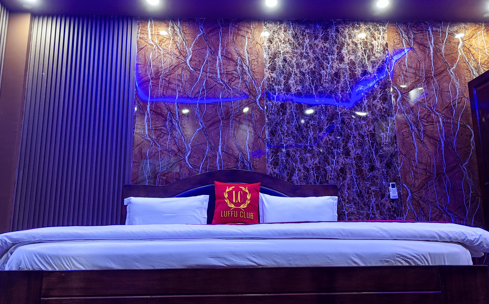 Wide view of a hotel bed against a dark, textured wall with recessed blue lighting - Luffu Club