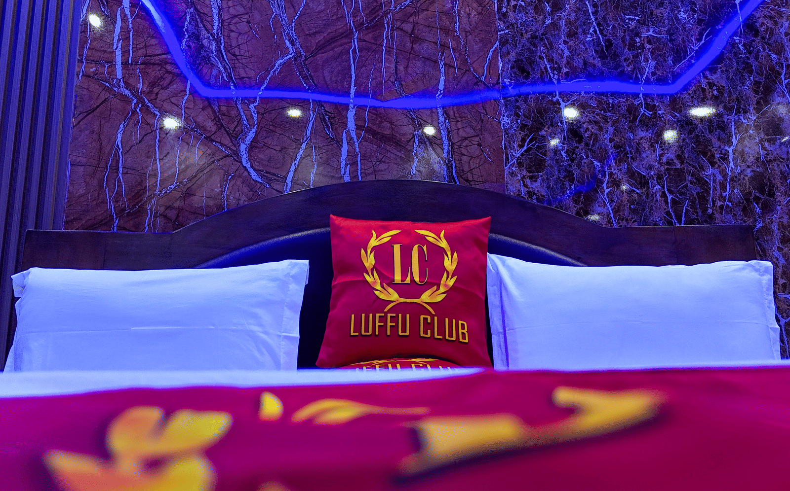 Close-up of a hotel bed with white pillows and a red cushion displaying a 'Luffu Club' logo - Luffu Club