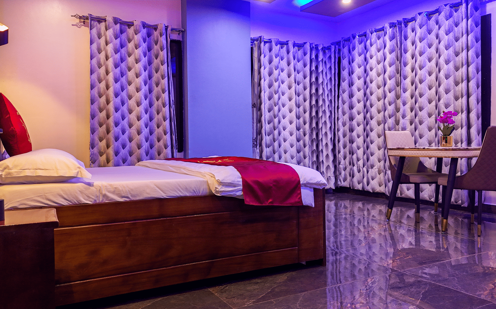 Interior view of a spacious room with a bed, small table and 2 chairs, patterned curtains, and vibrant purple lighting - Luffu Club