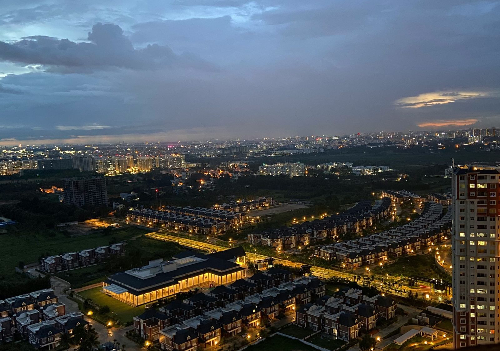An aerial view of a cityscape at dusk with lights, buildings, and a cloudy sky.