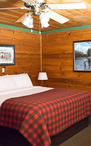 Overview of a Bedroom at Shaver Lake Village Hotel with wooden walls, plaid comforter, white pillows, framed artwork, and a ceiling fan.