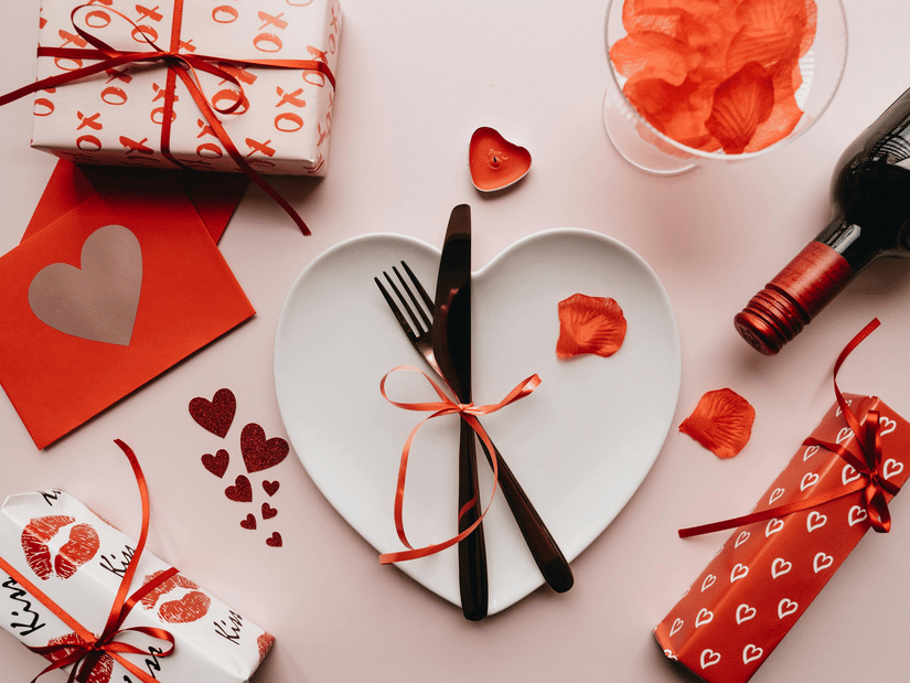 An image of a platter with cutlery tied with a ribbon, and around that there are heart shaped cards, gifts, and a wine bottle meant for valentine's day celebration