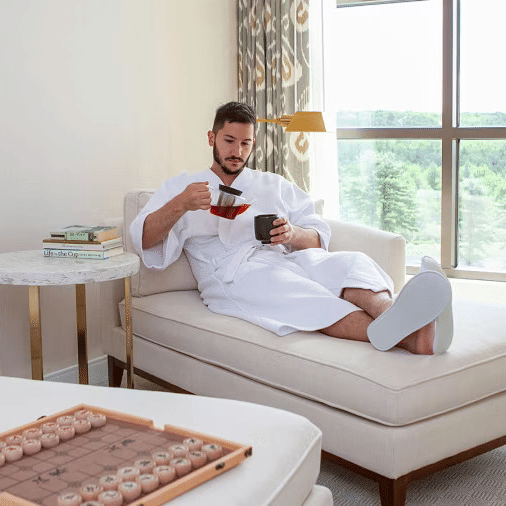 A man in a bathrobe on a couch at YO1 Longevity and Health Resorts pouring hot beverage from the kettle to a cup