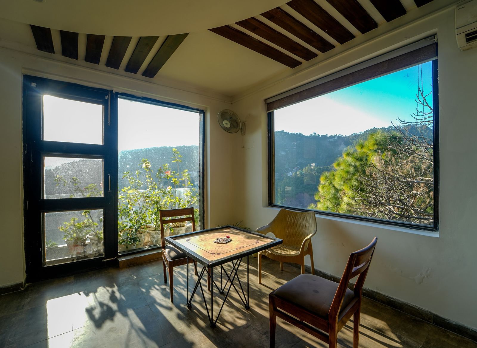 An image of the indoor game zone featuring a carrom board placed on a table with two chairs on either side and an a glass door and window overlooking the view - DLS Whispering Winds