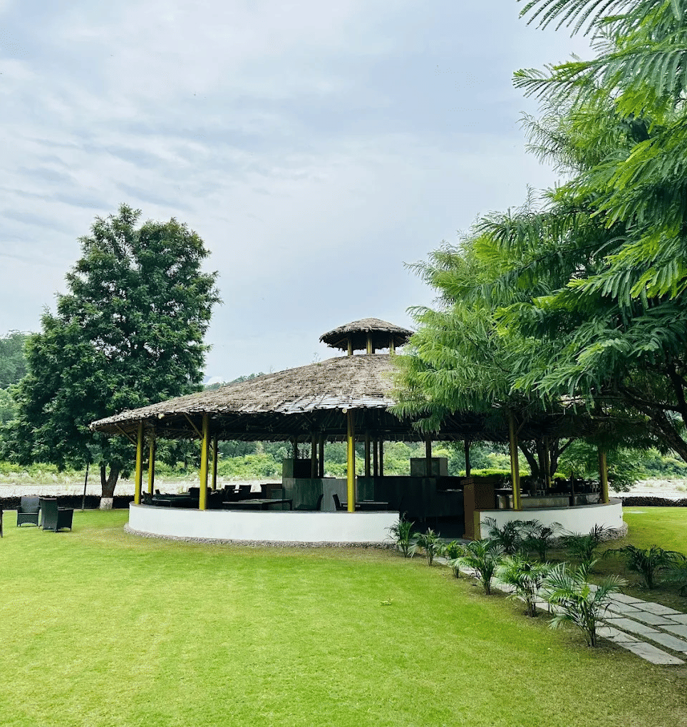 Green garden with a circular thatched-roof pavilion, outdoor seating, trees, and scenic hills in the background