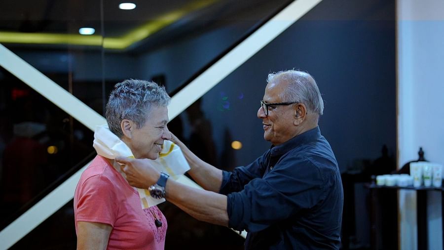 A man with glasses adjusts a white garment around a woman's neck in a salon or similar setting.