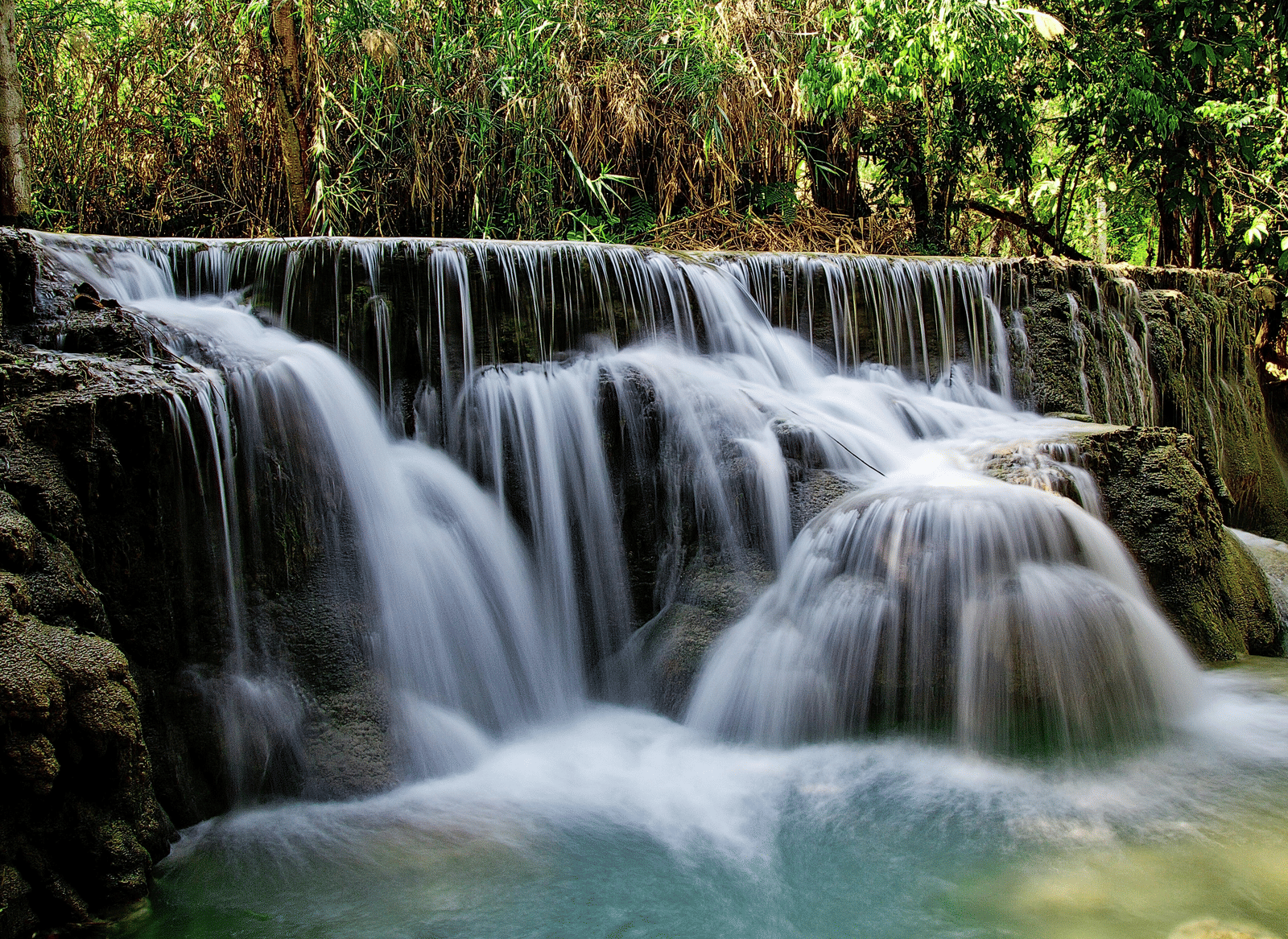 A wide, long-exposure shot of a multi-tiered waterfall flowing over smooth, light-coloured rocks.