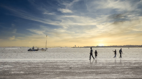 Several people walk along a wet sandy beach at sunset, with a boat anchored in the background.